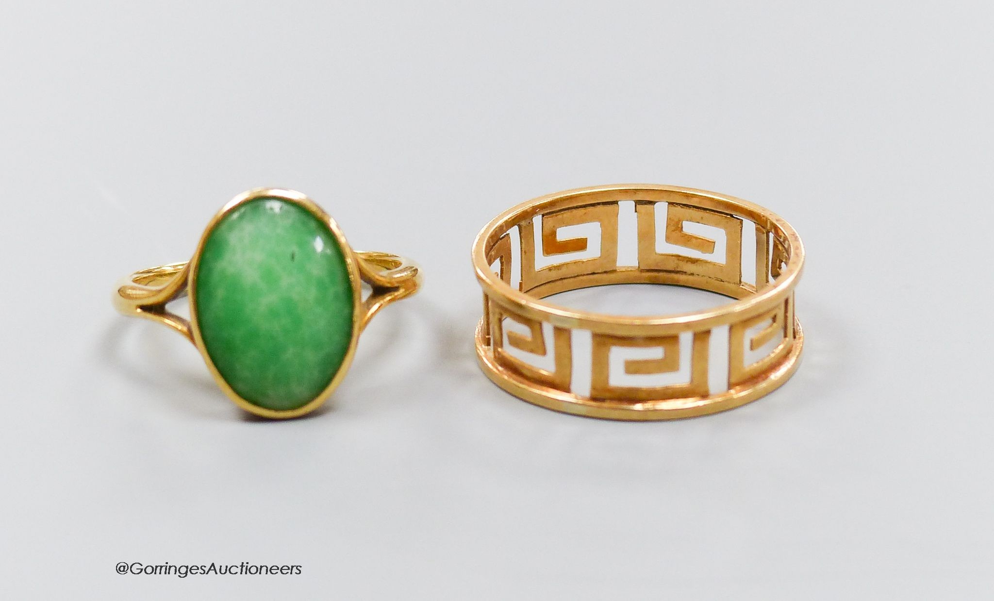 A yellow metal pierced Greek key pattern ring (testing as 18ct gold), size P, 2.2g and a similar ring set with an oval cabochon jade, size K, gross 3.2g. 5.3g